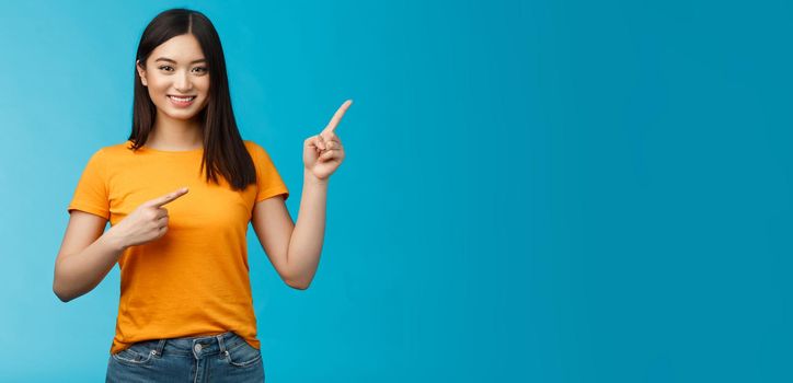 Friendly cheerful asian brunette girl inviting visit link interesting place, pointing upper right corner smiling joyfully camera, telling you about good promotion, stand blue background upbeat.