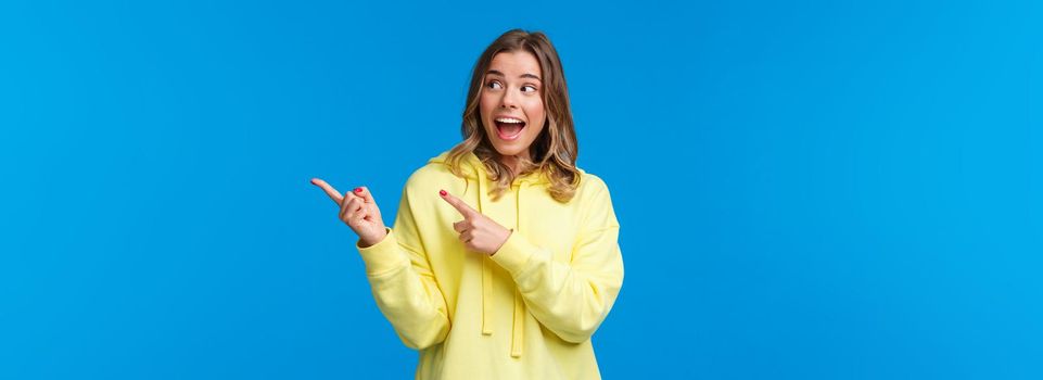 Excited happy, cheerful caucasian female with white beaming smile, wear yellow hoodie, react thrilled and amused to awesome news, cool banner, pointing fingers right, look away, blue background.