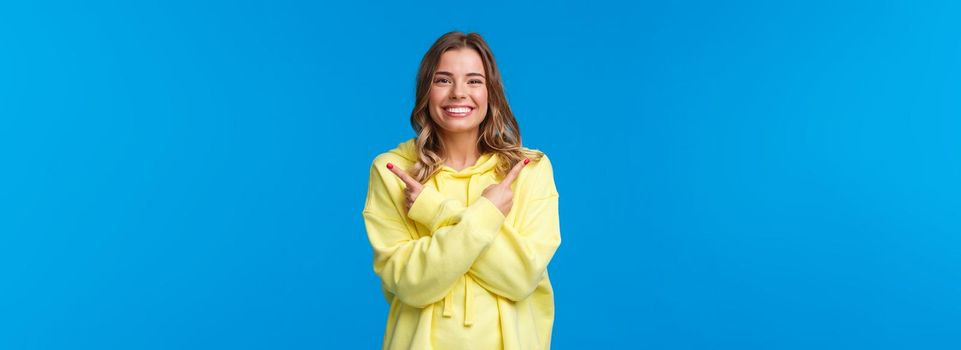 Confident pleasant, friendly-looking caucasian female showing two variants, give opportunity to choose what best, pointing fingers sideways and smiling camera, blue background.