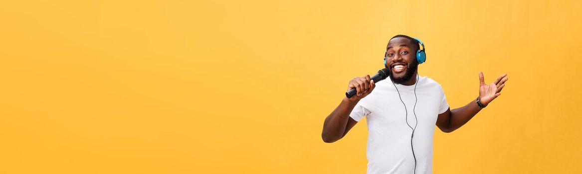 Portrait of cheerful positive chic. handsome african man holding microphone and having headphones on head listening music singing song enjoying weekend vacation isolated on yellow background.