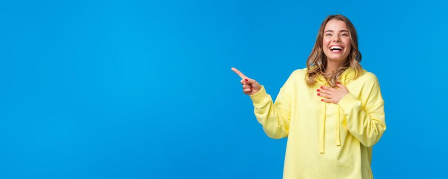 Happy and amused good-looking blond female in yellow hoodie, laughing out loud over something hilarious on upper left corner of blue background, smiling and looking entertained camera.