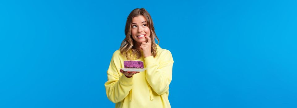 Celebration, party and lifestyle concept. Dreamy cute and silly blond girl thinking what to wish, biting finger and looking thoughtful up, smiling holding b-day cake, blue background.