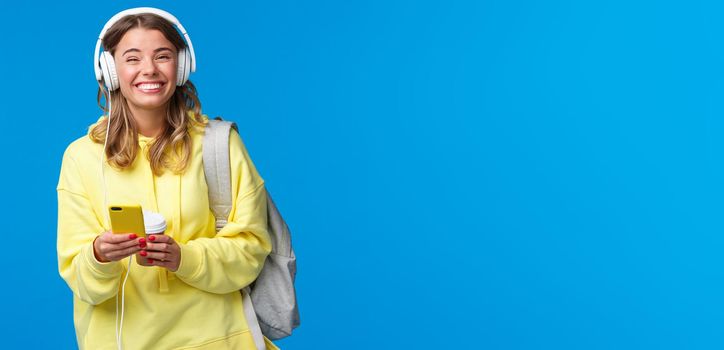 Cheerful pretty young female student with backpack, listening favorite playlist in headphones, laughing and smiling at camera carefree, hold mobile phone, stand blue background.