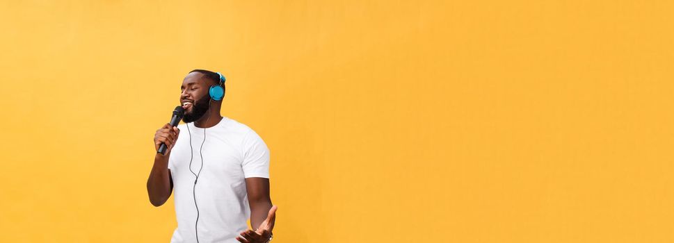 Portrait of cheerful positive chic. handsome african man holding microphone and having headphones on head listening music singing song enjoying weekend vacation isolated on yellow background.