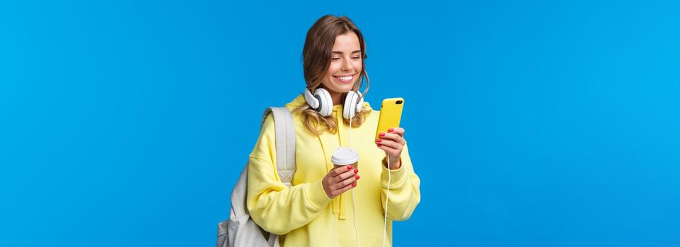 Girl using mobile phone to contact friend after college, texting as walking along street with backpack, take-away coffee cup and headphones, smiling joyfully smartphone display.