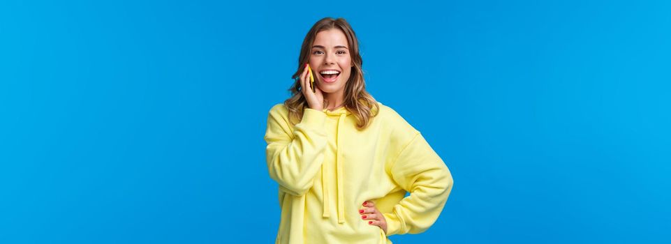 Carefree good-looking european blond girl in yellow hoodie lively talking on mobile phone, smiling camera as booking table for friends in bar, order food delivery, blue background.