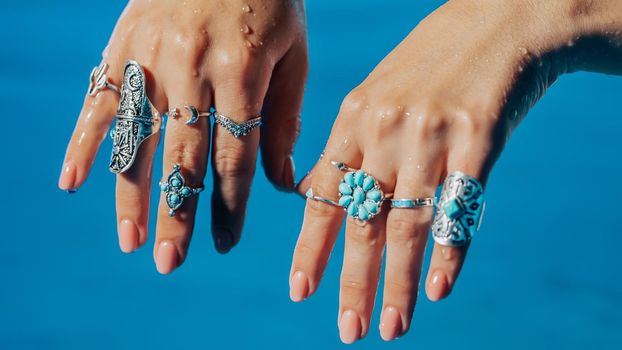 Beautiful gypsy woman demonstrates boho jewelry, rings with turquoise stones on hands. Girl in white dress showing accessories in blue water. Femininity, trend, hippie style concept.High quality photo