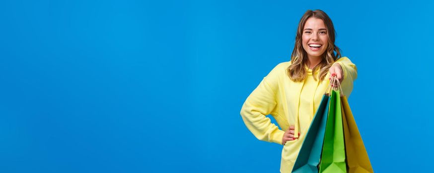 Cheerful blond girl giving you shopping bags and smiling happy, buying presents for family holiday, buying lots of staff in store and asking carry her packages, stand blue background.