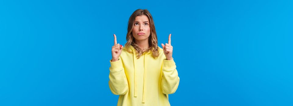 Jealous and gloomy cute blond female student feel regret not having something she desires, sobbing frowning sad as looking pointing up, standing over blue background.