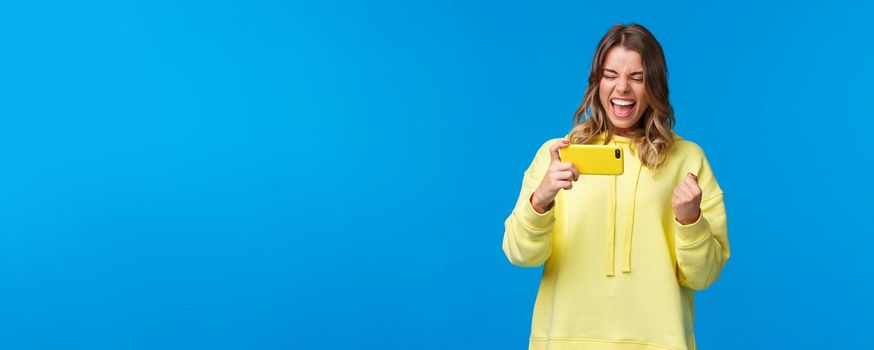 Satisfied and pleased triumphing young girl watching online live stream of game in internet using mobile phone, winning, rooting for team scored goal, saying yes delighted, blue background.