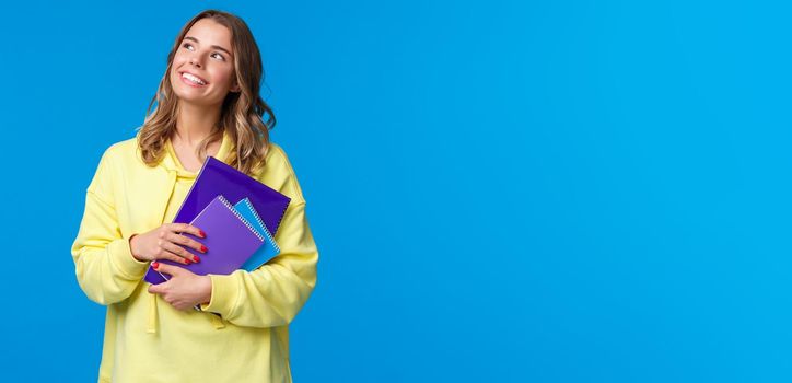 Back to school. Cute blond smiling european female student carry notebooks and learning material, look dreamy upper left corner smiling, thinking what she grab for lunch, blue background.