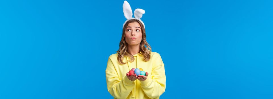 Thoughtful cute caucasian girl in rabbit ears thinking, looking up pondering and dreamy, holding Easter eggs, decide where hide it during traditional holiday game, blue background.