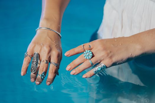 Beautiful gypsy woman demonstrates boho jewelry, rings with turquoise stones on hands. Girl in white dress showing accessories in blue water. Femininity, trend, hippie style concept.High quality photo