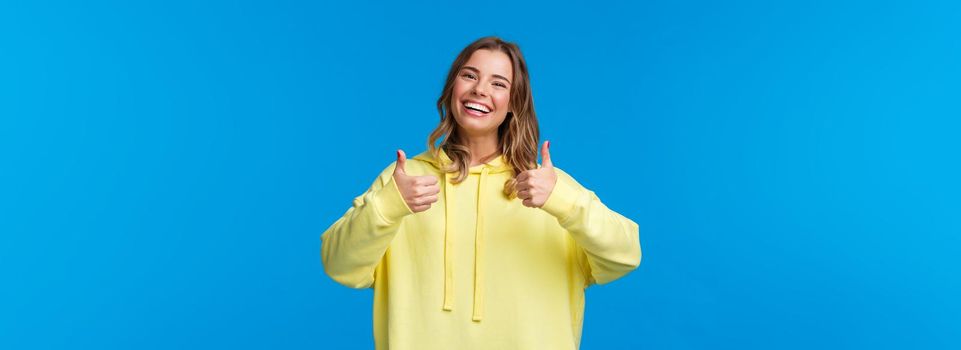 Sounds good Im in. Cheerful and optimistic good-looking young female blonde in yellow hoodie, show thumbs-up in approval, smiling nod acceptingly, like idea, blue background.