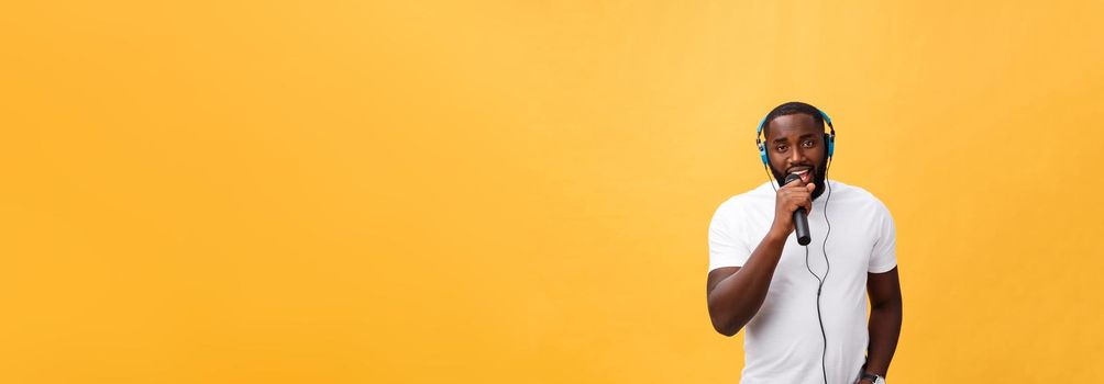 Portrait of cheerful positive chic. handsome african man holding microphone and having headphones on head listening music singing song enjoying weekend vacation isolated on yellow background.