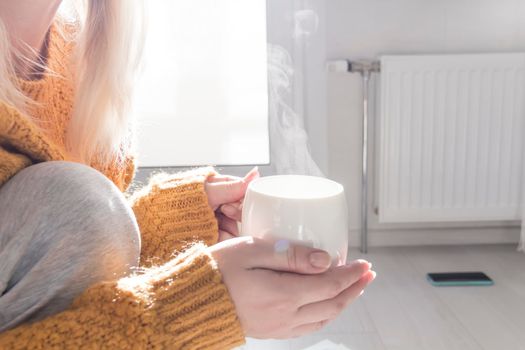 A young girl in a yellow sweater and hat is drinking hot tea, near a heater with a thermostat..