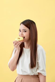 Portrait of a lovely young asian woman eating donuts isolated over yellow background