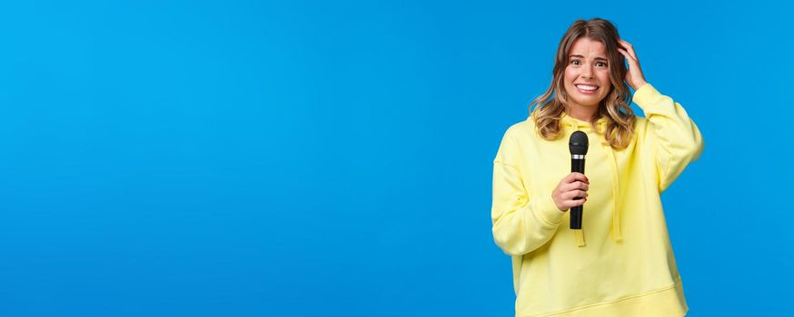 Embarrassed girl holding microphone awkward smiling and scratch head as singing song on karaoke with friends, shy perform in front of audience, standing blue background.