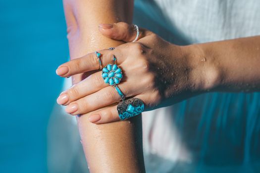 Beautiful gypsy woman demonstrates boho jewelry, rings with turquoise stones on hands. Girl in white dress showing accessories in blue water. Femininity, trend, hippie style concept.High quality photo