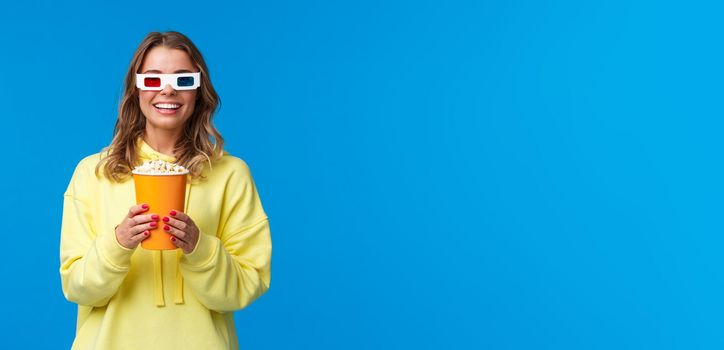 Lets watch movie. Excited cute young blond girl in 3d glasses, holding popcorn and smiling amused as enjoying premiere of film in cinema, standing entertained over blue background.