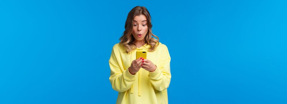 Surprised and intrigued good-looking blond female seeing something interesting online, browsing internet searching outfit in clothes store, using cool new application, hold smartphone.