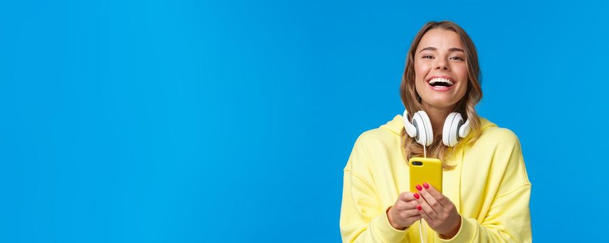 Close-up portrait joyful good-looking blond girl in yellow hoodie and white headphones, using mobile phone, writing in her blog, sharing news with social media, messaging and laughing camera.