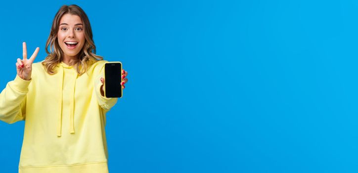 Optimistic cute blond european girl with short haircut, yellow hoodie, showing kawaii peace gesture and mobile phone display as using photo filter to edit and post pic online, blue background.