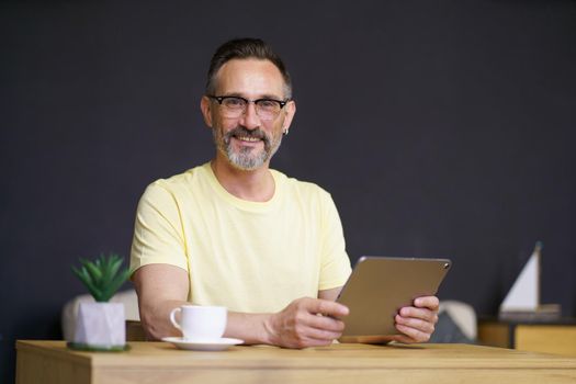 Working from home middle aged man use digital tablet sitting at home office desk wearing casual with black wall on background. Handsome freelancer mature man hold tablet pc working from home.