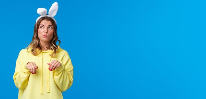 Thoughtful and cute funny blond girl acting like Easter rabbit with fake ears and paws gesture, pouting looking upper left corner mimicking bunny animal, stand blue background.