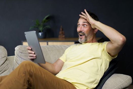 Middle aged handsome man use digital tablet to communicate via video call or reading news with shock facial expression sitting on the sofa in home interior wearing casual. Freelancer, work from home.