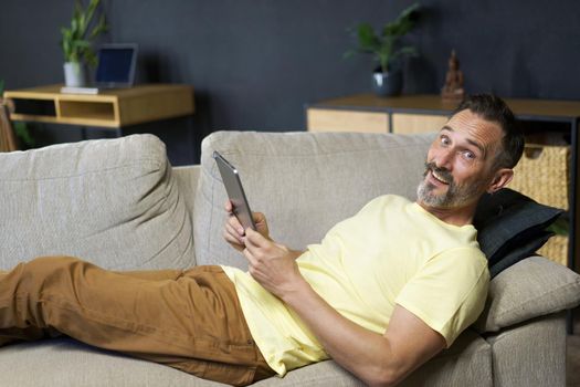 Laying on the couch middle aged freelancer man use digital tablet to read social media or working offline from home wearing casual. Relaxed freelancer man laying on sofa making business uses gadget.