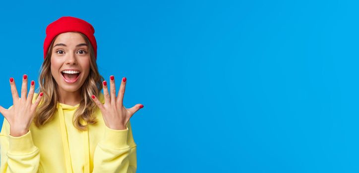 Portrait of amused good-looking blonde hipster girl smiling joyfully, explain something showing number ten, tenth with hands and look camera upbeat, order dozen, stand blue background.