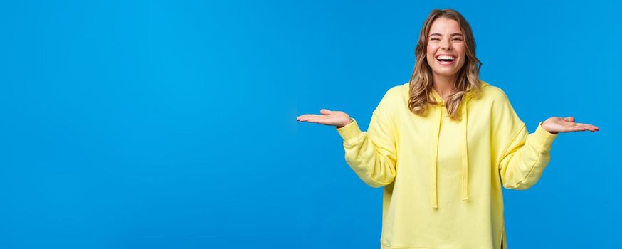 Happy carefree good-looking woman, blonde short haircut, holding both hands raised sideways as if holding two products, give you choice and laughing, introuce two products, blue background.