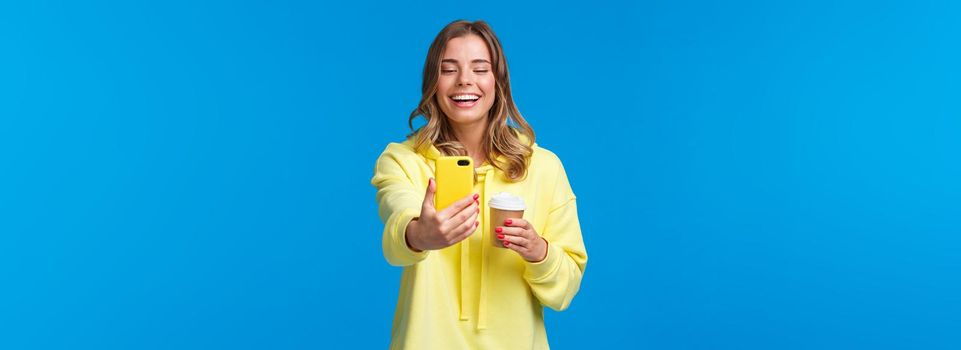 Cheerful smiling blond caucasian female blogger record video or take selfie on her smartphone, laughing and grinning as holding take-away cup of coffee, stand blue background.