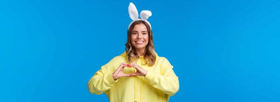 Holidays, traditions and celebration concept. Waist-up portrait of charismatic cute blond girl in rabbit ears, congratulate everyone with Easter holiday, show heart gesture and smiling.