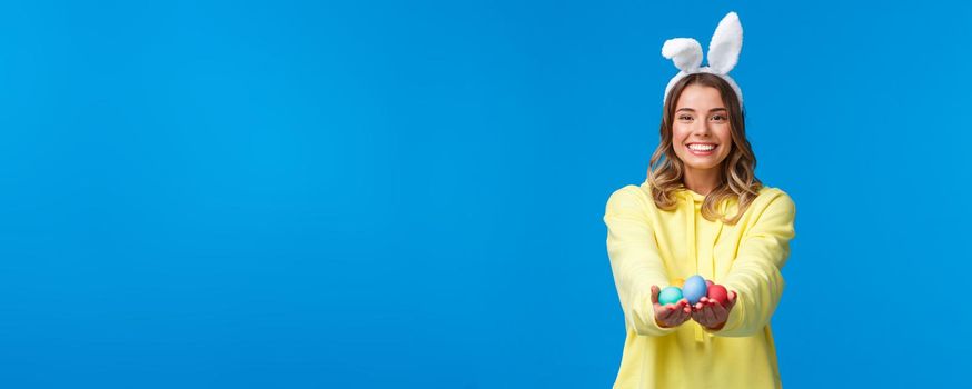 Holidays, people and fun concept. Charismatic cute blond girl holding coloured Easter eggs, wearing bunny ears and smiling joyfully, congratulate family with holy day, blue background.