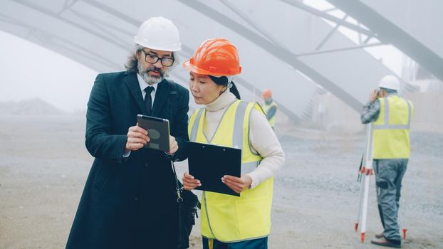 Young Asian woman engineer is talking to mature Caucasian man investor discussing construction project reading papers and using tablet outdoors
