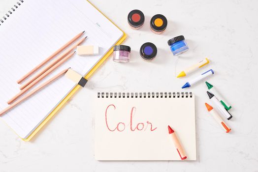 Bright multicolored pencils, crayons and watercolors bottles with color letters next to a sketchbook on white table