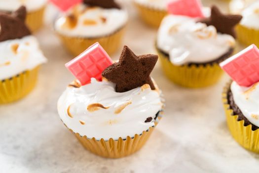 Gourmet s'mores cupcakes with meringue frosting and garnished with star-shaped chocolate graham cracker and a mini pink chocolate bar.