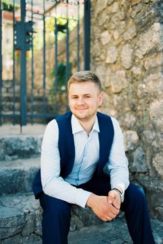 Smiling groom sits on stone steps in front of a forged gate. High quality photo