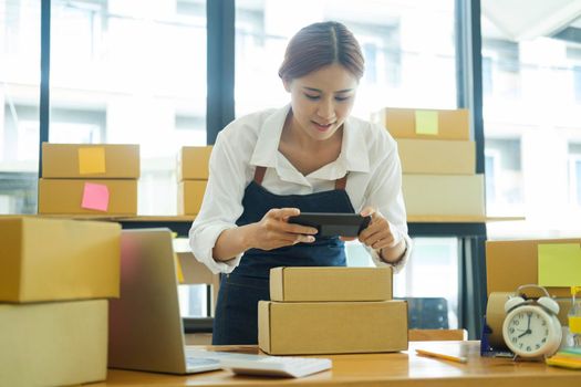 Asian female online business owner or online store owner using cellphone taking picture and checking parcel box, scanning retail package parcel barcode on shipping box preparing for delivery and shipment.