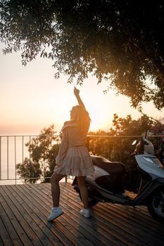 A young beautiful woman in summer clothes stands on the background of an orange sunset near the city moped. selective focus