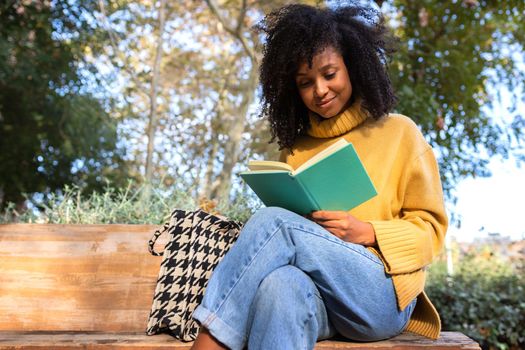 Happy young African American woman sit on a park bench reading a book. Copy space. Lifestyle concept.