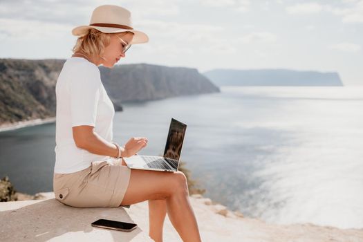 Successful business woman in yellow hat working on laptop by the sea. Pretty lady typing on computer at summer day outdoors. Freelance, travel and holidays concept.