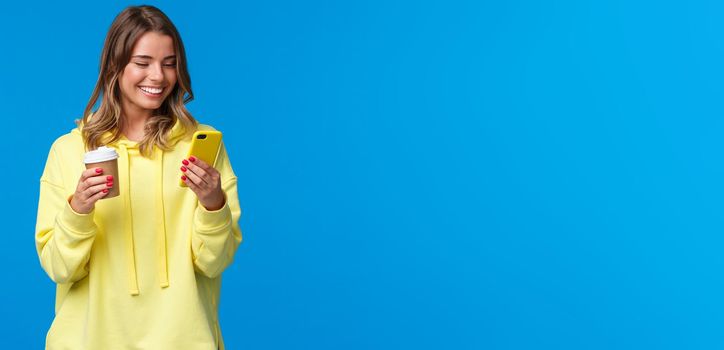 Carefree joyful european female in yellow hoodie, holding take-away coffee and browsing internet in mobile phone, texting friend laughing over funny video or meme, blue background.