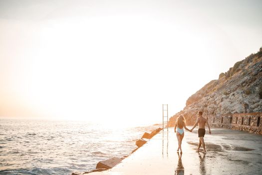 A loving couple walks along the beach by the sea. Young family at sunset by the Mediterranean Sea. Vacation concept. A woman in a swimsuit and a man in shorts at sunset by the sea. Selective focus.