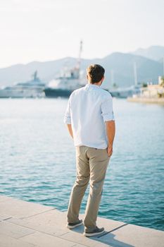 Young man stands on the pier and looks at the moored yachts. Back view. High quality photo