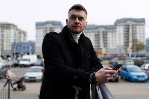 portrait of a brutal young businessman in a stylish black coat with a phone in his hands on the background of the city.