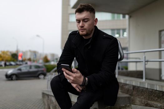 brutal young man in a black coat with a mobile phone on the background of the office center.