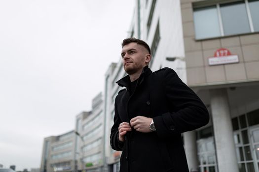 stylish brutal young man on the background of a city building in a black coat.